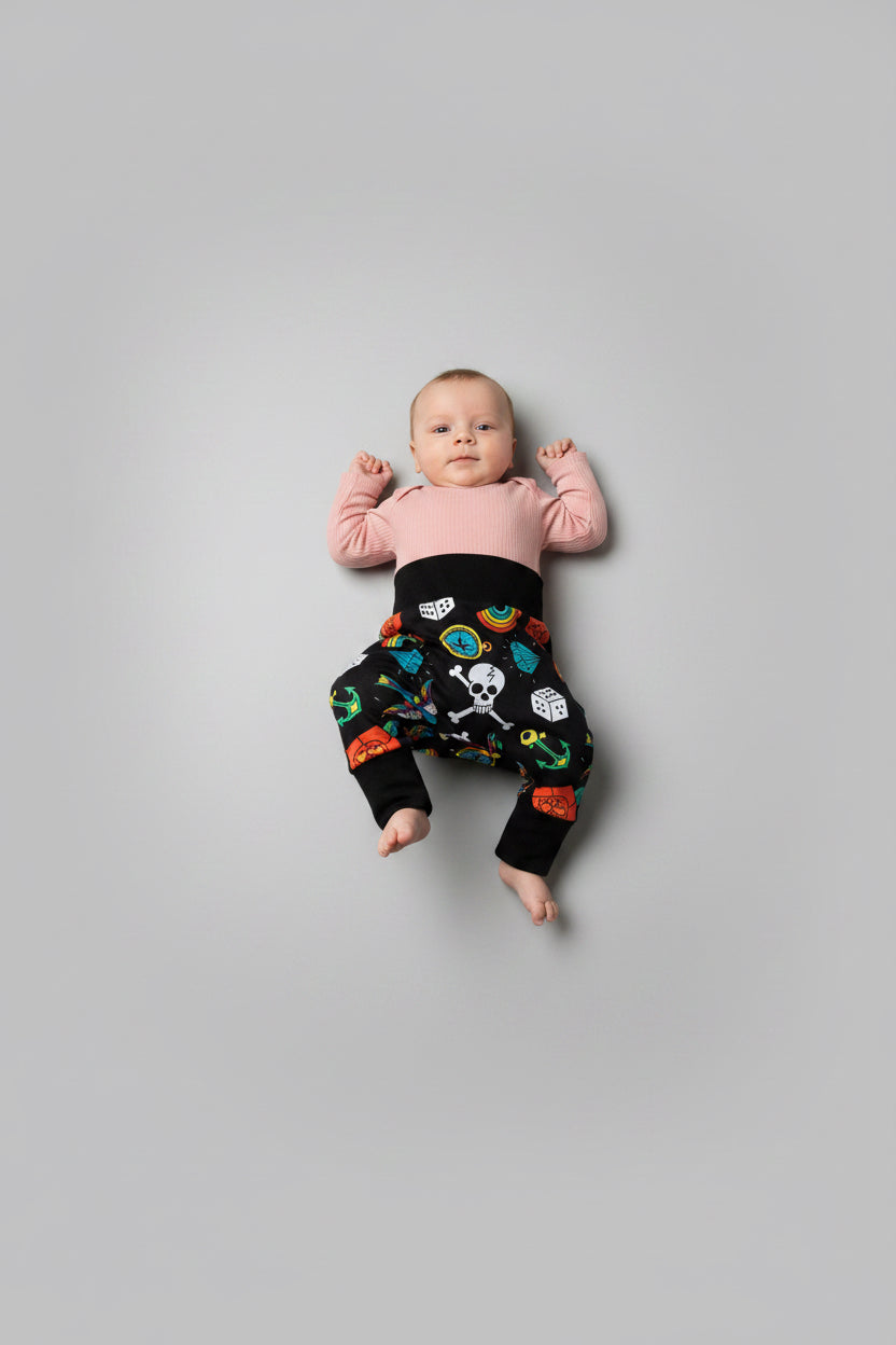 A baby lying on their back against a solid gray background, wearing a light pink long-sleeve top and black pants with a colorful tattoo pattern.