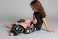 Two children sitting on a light gray studio floor. The older child wears a black graphic T-shirt and colorful tattoo patterned leggings, while the younger child wears a light pink long-sleeve top and matching tattoo print patterned leggings.