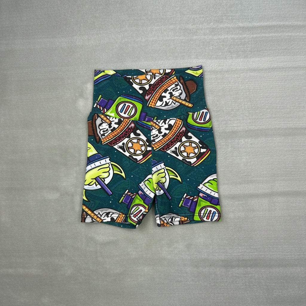 A pair of kids' shorts featuring a colorful Buzz Lightyear space-themed print on a dark teal background, laid flat on a gray textured surface.